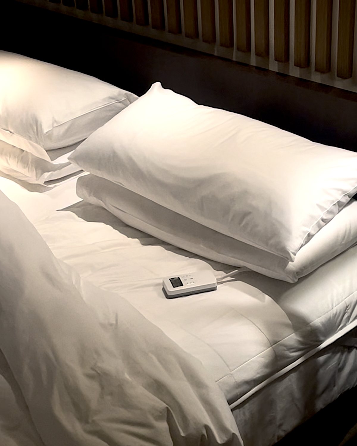 Litual on a hotel bed at night — one pool of light on the pillows, the quilted topper under white linen, its controller resting on the bed