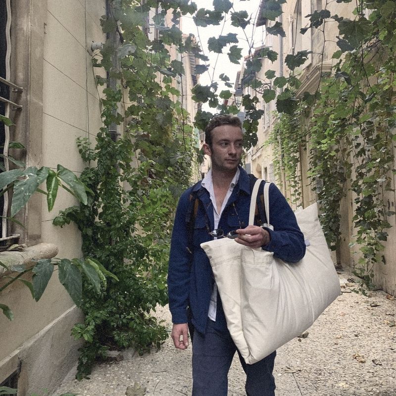 A man walking down a vine-covered lane with a large cream Litual bag over his shoulder