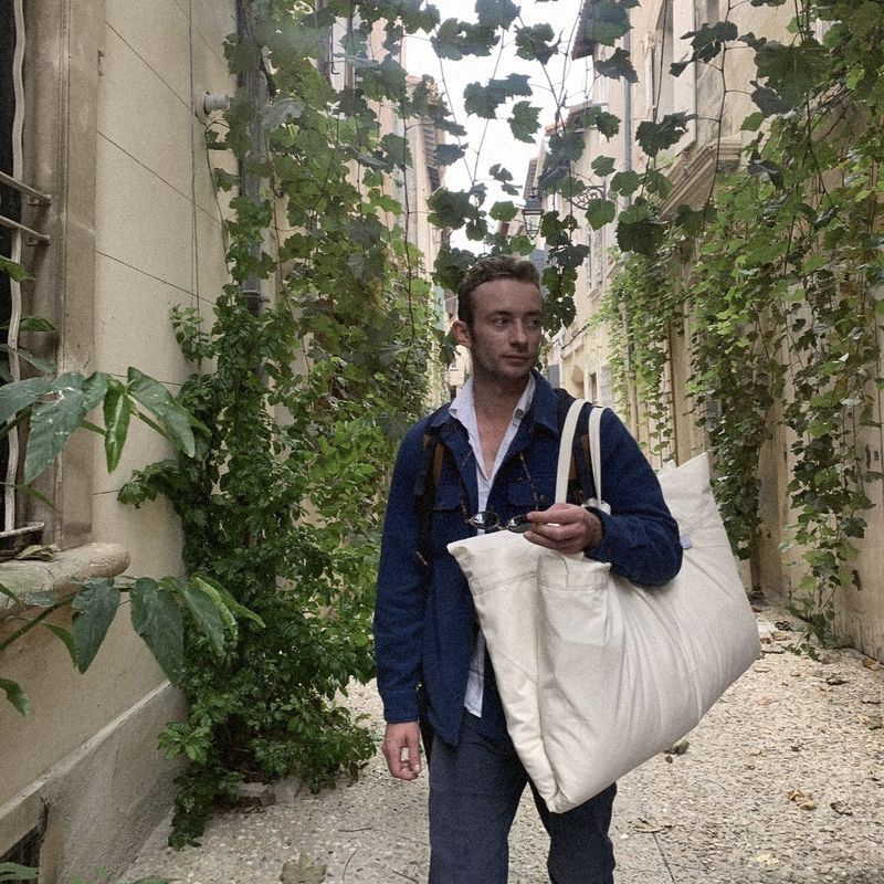 A man walking down a vine-covered lane with a large cream Litual bag over his shoulder