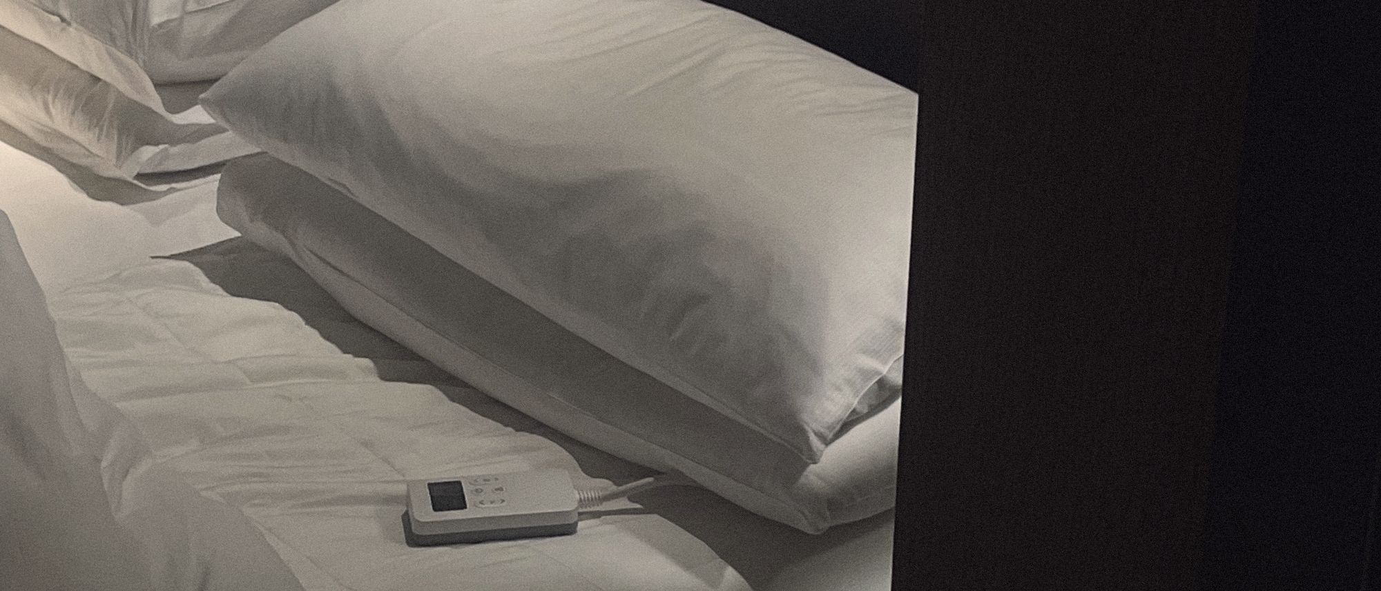 A hotel bed at night — pillows lit by a single lamp, the Litual controller resting on the sheet