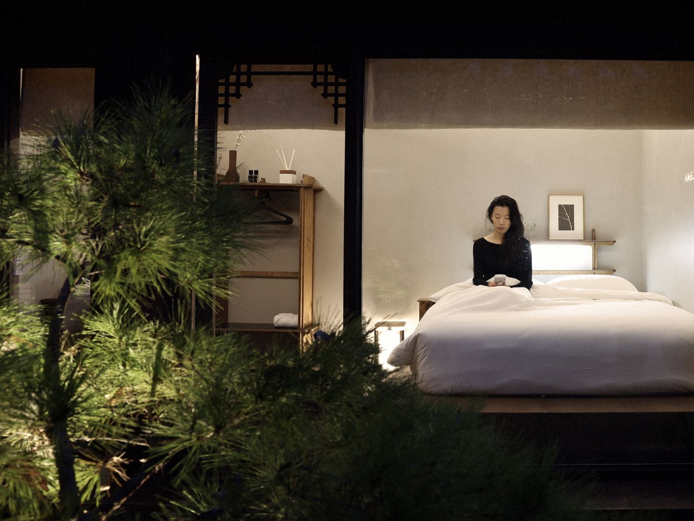 A hanok at night — through the window, a woman sits up in a white bed, the room lit warm from within