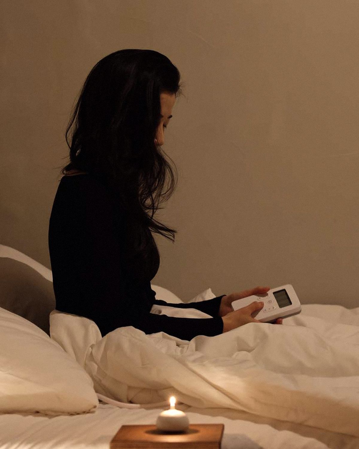 A woman sitting up in bed at night, hair down, setting the Litual controller by candlelight
