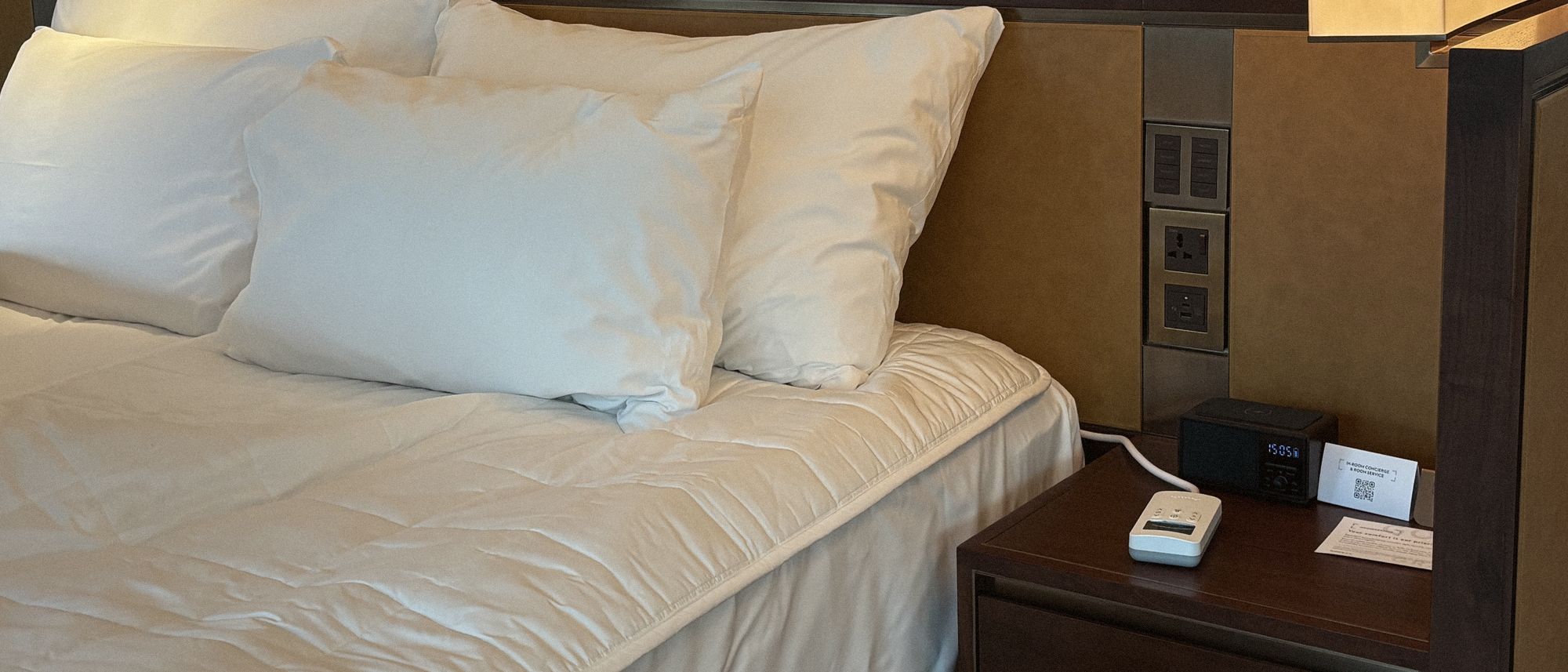 A hotel bed under warm headboard light, the quilted Litual on the mattress and its controller on the bedside table