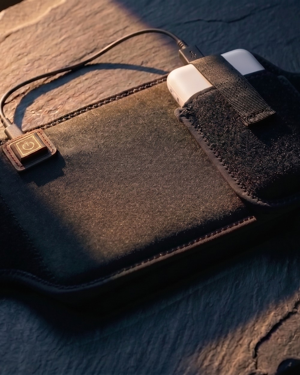 The Poom wrap on dark slate at dusk — the matte black band, its power button lit, and a power bank held in the pocket by its strap