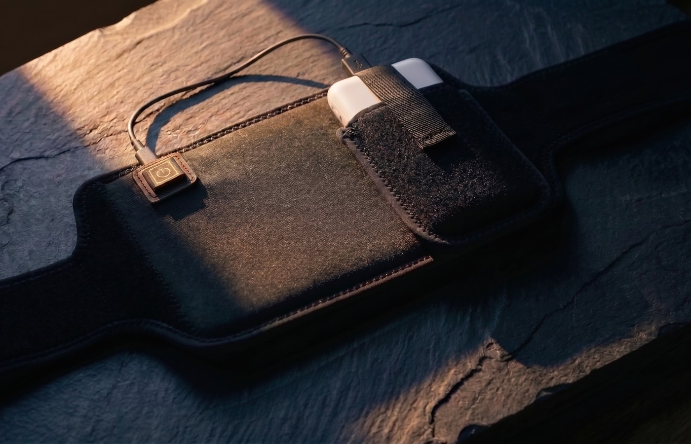 The Poom wrap open on dark slate in low evening light — the matte black band, its power button lit, and a power bank held in the pocket by its strap